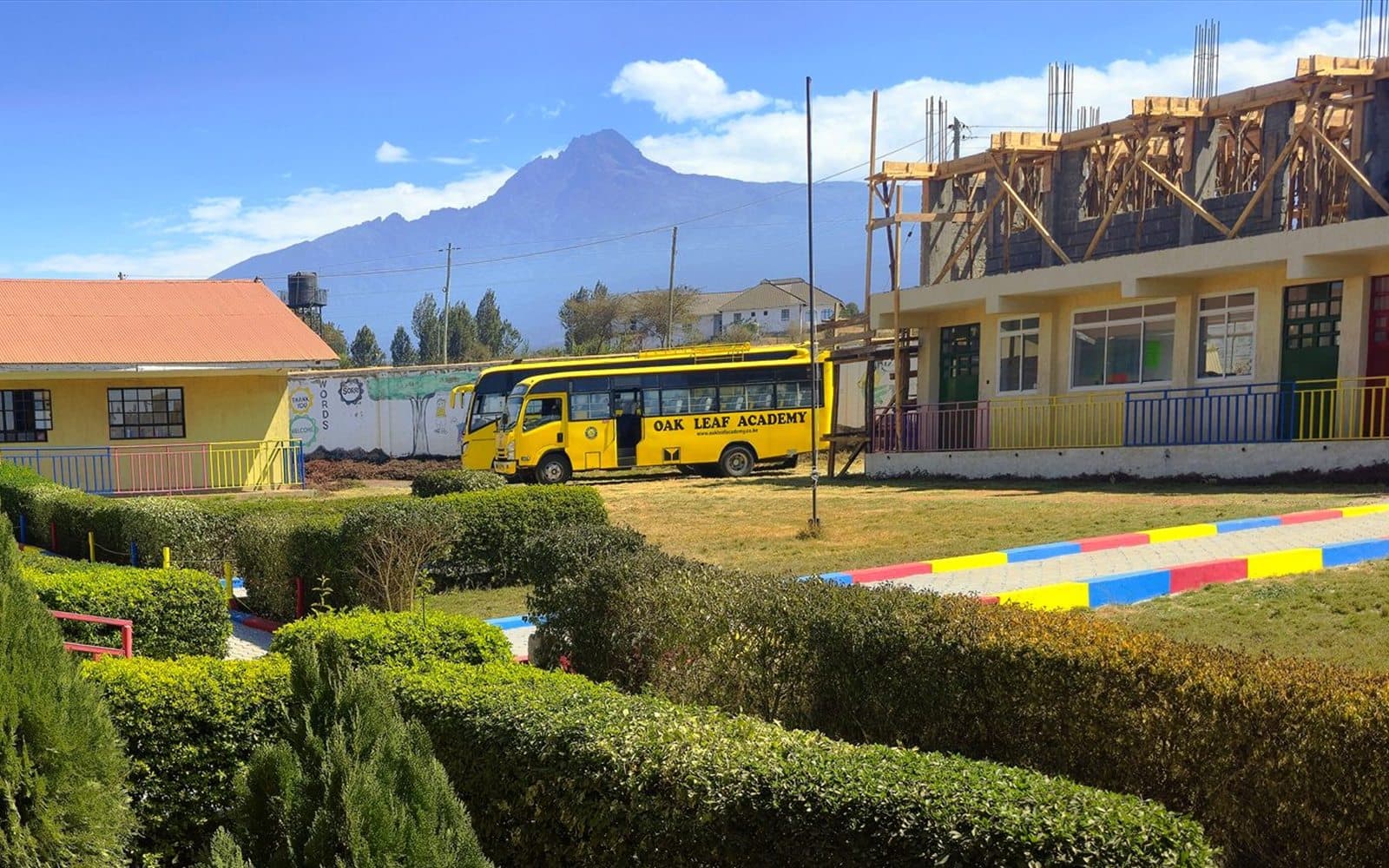 The yellow Oak Leaf Academy school bus parked on the campus lawn beside the classroom block, with Mount Kilimanjaro rising behind it.